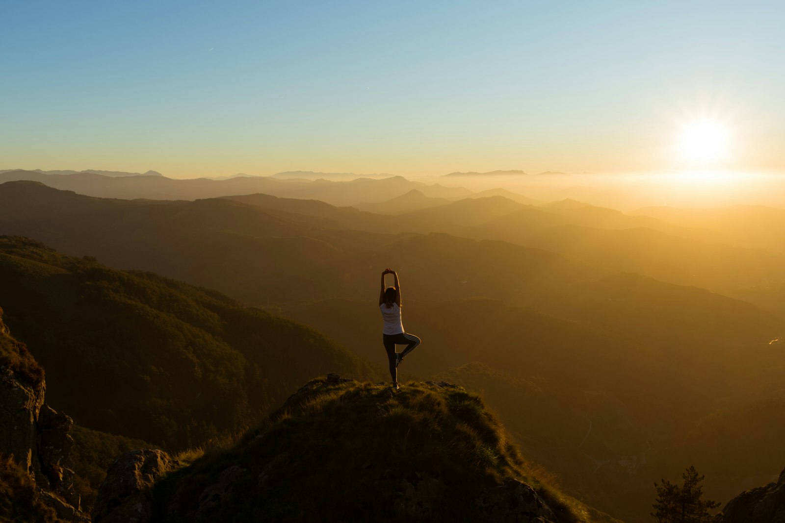 A woman balances in tree pose on a grassy summit as the sun rises over layered mountain ridges.