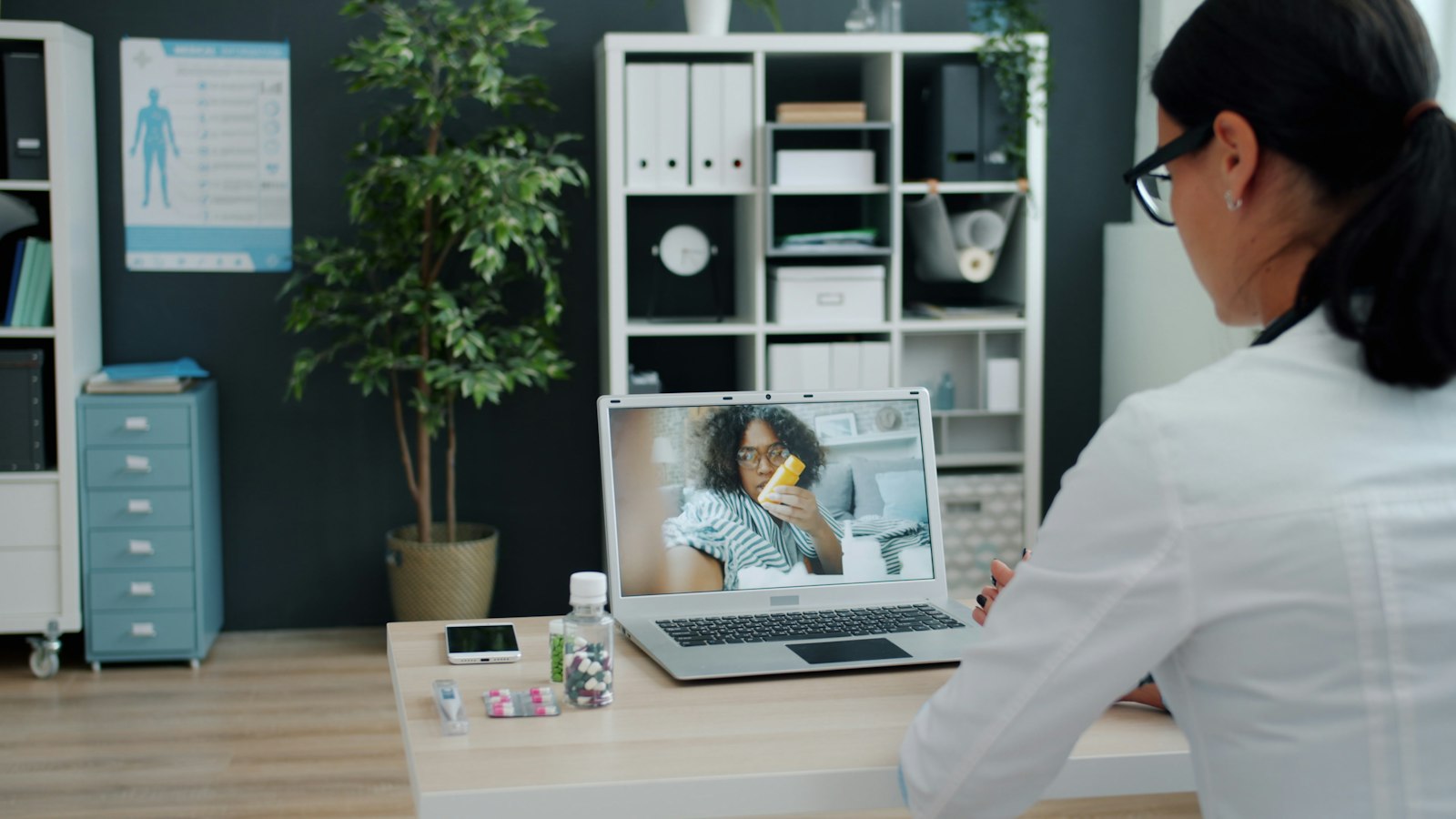 A provider in a bright office talks by laptop video with a patient who is resting at home under a blanket.